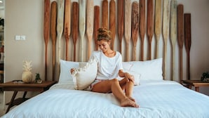 A bed with a white sheet, a wooden headboard with wooden paddles, and a person sitting on the bed. - Villa Bloom 2 Bedroom (Seminyak)