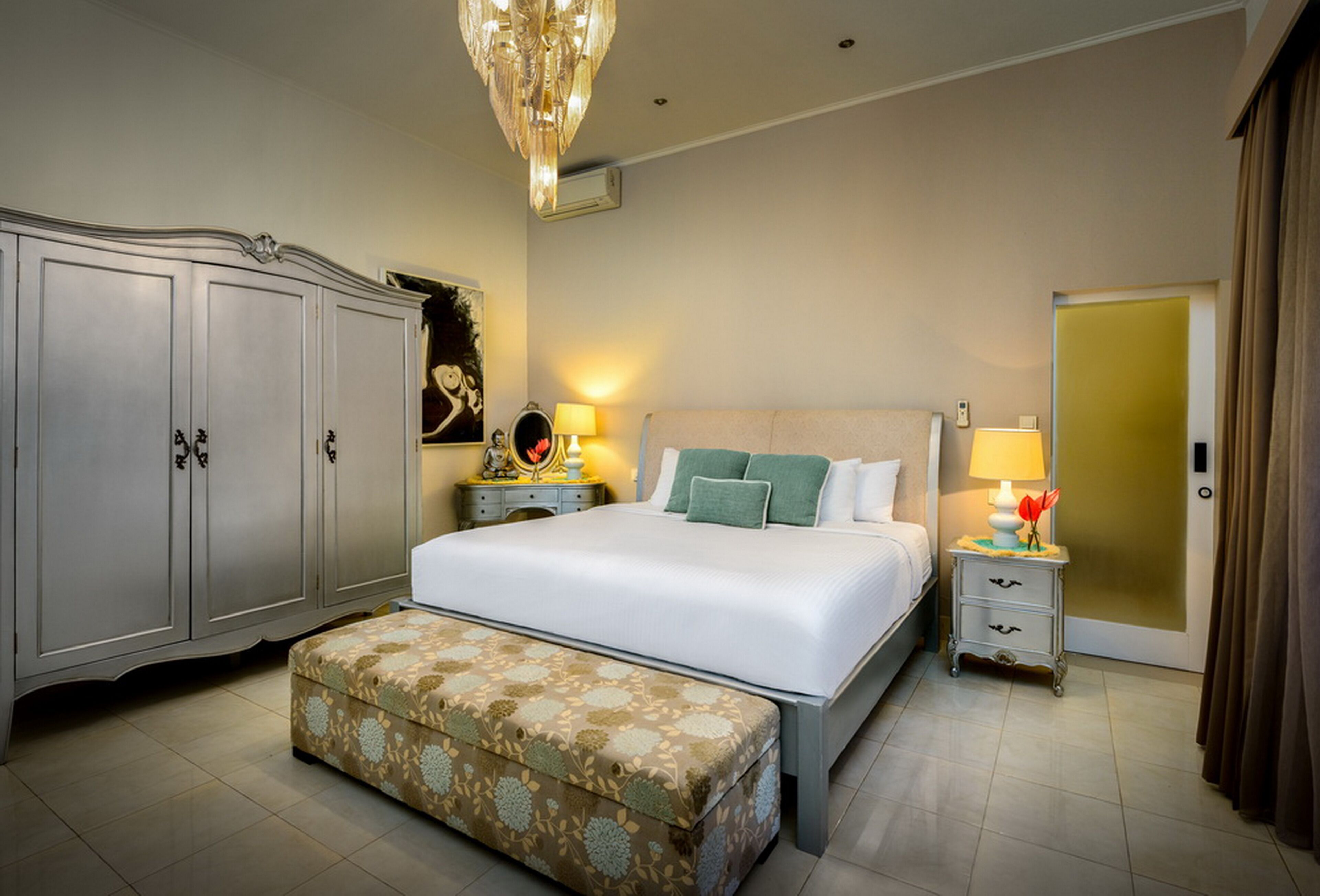 A well-lit bedroom with a large bed, a chest of drawers, a bedside table with a lamp, and a door with curtains.