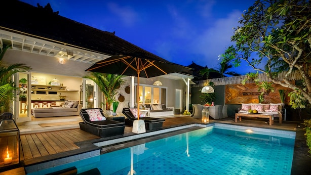 A poolside area with lounge chairs, a dining area, and a kitchen visible through an open door. - Villa Bloom 2,5 Bedroom (Seminyak)