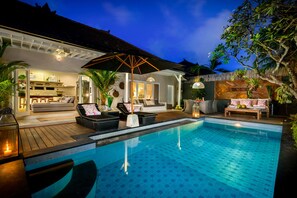 A poolside area with lounge chairs, a dining area, and a kitchen visible through an open door. - Villa Bloom 2,5 Bedroom (Seminyak)