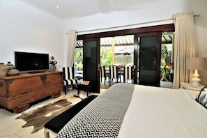 A modern hotel room with a large bed, a television, a wooden dresser, and a sliding glass door leading to an outdoor seating area.