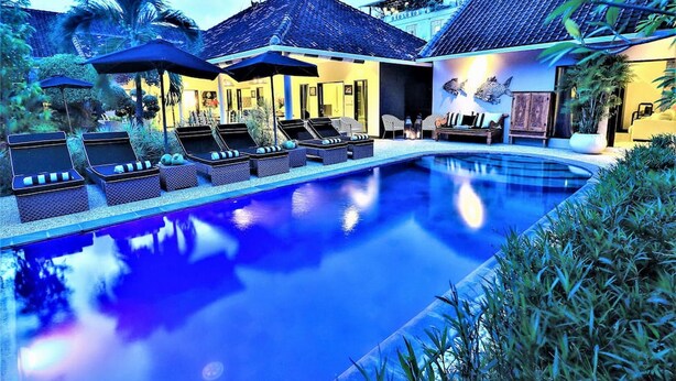 A swimming pool with lounge chairs and umbrellas, surrounded by greenery and a building with a tiled roof. - Villa Bloom 4 Bedroom (Seminyak)