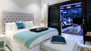 A modern bedroom with a tufted headboard, a large bed with a blue and white color scheme, a bedside table with a lamp, and a view of a balcony with outdoor furniture. - Villa Bloom 4 Bedroom (Seminyak)
