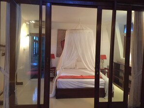A bedroom with a canopy bed, wooden headboard, and a nightstand with a lamp. - Villa Hangat (Seminyak)