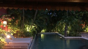 Zona de piscina con una silla de descanso y una lámpara, rodeada de plantas tropicales y techo de madera.