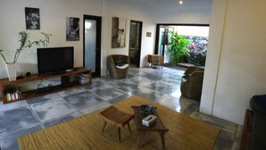 Sala de estar com televisão, estante, mesa de centro e duas cadeiras. Uma porta dá acesso a uma varanda com plantas.