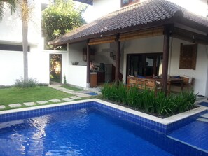 A residential pool area with a tiled deck, a covered seating area, and a well-maintained lawn.