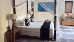 Uma cama com dossel e cortina, com edredom branco, almofadas preto e branco e manta preta. Tem uma mesa de cabeceira de madeira com uma luminária e um controle remoto. Um banquinho de vime com almofadas é visível ao fundo.