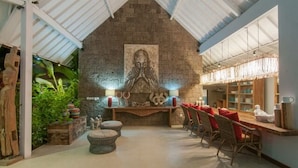 Uma área de refeições acolhedora com uma parede de pedra, uma estátua central de Buda e uma longa mesa de madeira com almofadas vermelhas.