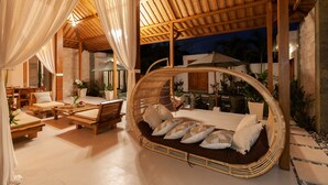 A cozy outdoor seating area with a wicker swing chair, cushioned sofas, and a wooden table. - Villa Kubu Joglo (Seminyak)
