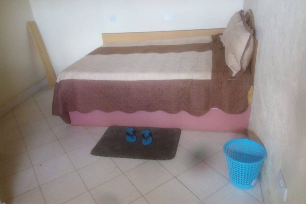 A small, single-bed room with a wooden headboard, a brown and pink bedspread, and a pair of blue slippers on a small rug. A blue laundry basket is placed against the wall.