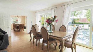 Een eetruimte met een houten tafel en stoelen, een bloemenvaas en grote ramen met gordijnen.