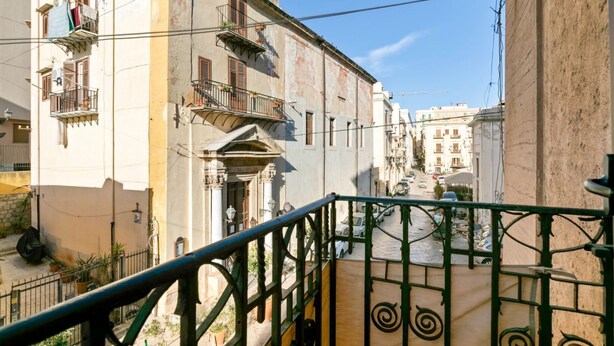 A narrow street with buildings, a balcony with a green railing, and a clear blue sky.