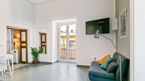 A living room with a sofa, a TV mounted on the wall, a door leading to a balcony, and a small table with a lamp.