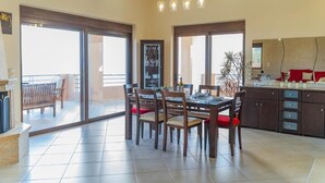 Een ruime eetruimte met een grote tafel en stoelen, een keuken met kastjes en een aanrecht, en grote schuiframen die leiden naar een buitenbalkon.