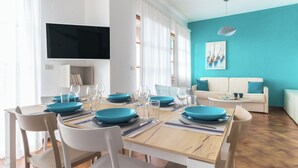 Uma sala de jantar com uma mesa de madeira posta para quatro pessoas, rodeada por cadeiras brancas. Há uma televisão na parede e um sofá com um almofada verde-água na sala de estar.