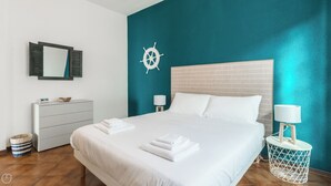 Quarto de hotel com uma cama, penteadeira, espelho, decoração de roda de navio e uma pequena mesa com abajur.