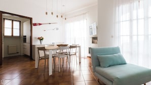 Uma cozinha moderna com armários brancos, uma mesa de jantar com cadeiras, um sofá azul e uma janela com cortinas leves.