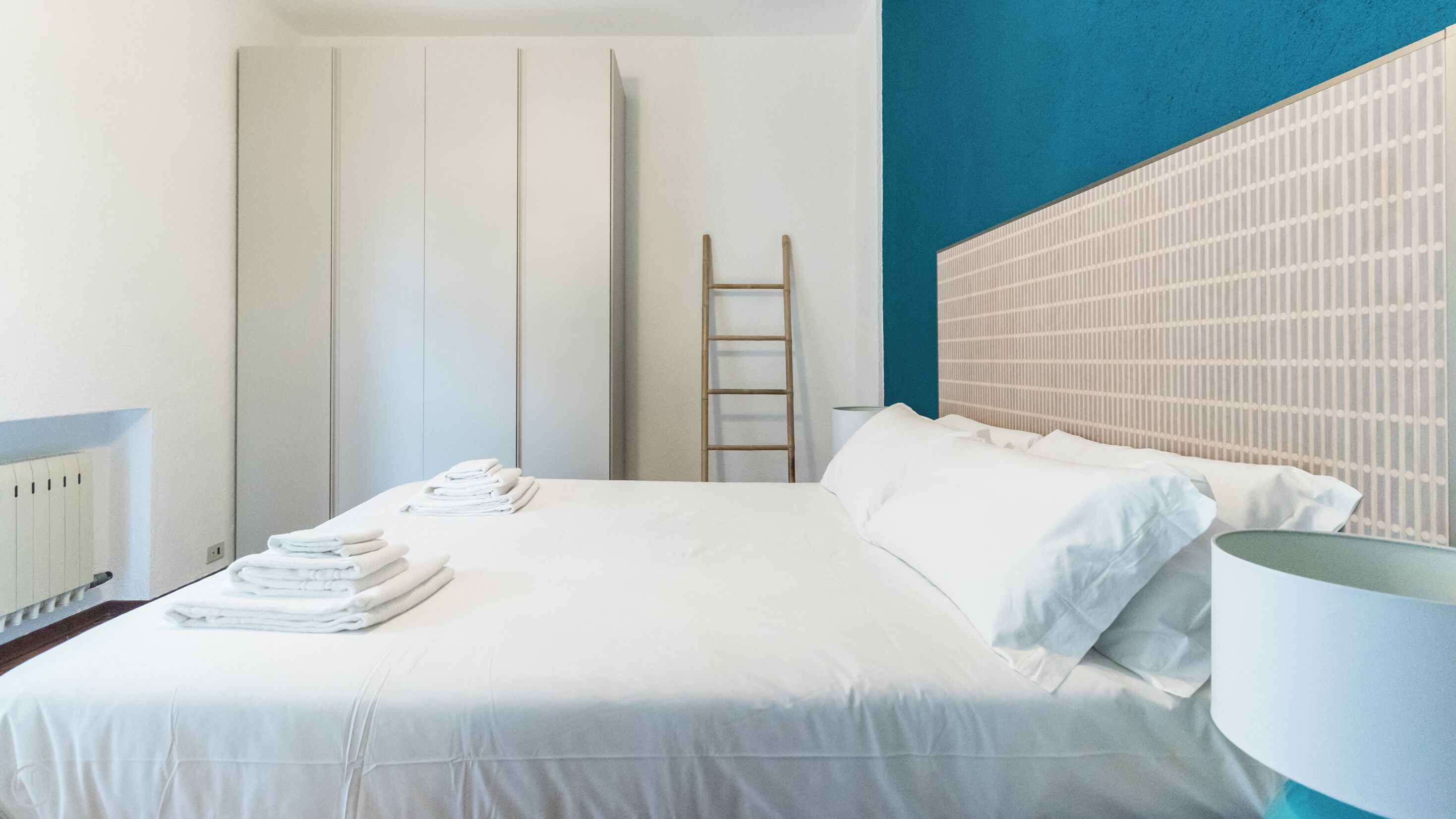 Una camera d'albergo con un letto rifatto, un set di asciugamani, una scala di legno e una parete con motivo a griglia.