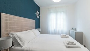 Una habitación de hotel con una cama grande, un cabecero con diseño de cuadrícula, un adorno de timón de barco en la pared, una lámpara de noche y una ventana con cortinas traslúcidas.