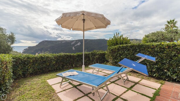 Un patio con dos tumbonas azules y un paraguas color beige, rodeado de setos y vistas a un paisaje montañoso.