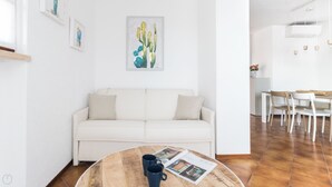 Um sofá branco com duas almofadas, uma mesa redonda de madeira com duas canecas azul-escuras e uma revista, e um quadro de cactos na parede.