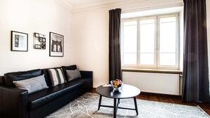 A modern living room with a black sofa, a round coffee table, and a geometric patterned rug. There are three framed pictures on the wall and a window with curtains. - Powisle Superior Apartment (Warsaw)