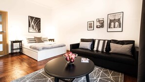 A modern living room with a bed, a black sofa, and a coffee table with apples. There are framed pictures on the wall, including one of the Brooklyn Bridge. - Powisle Superior Apartment (Warsaw)