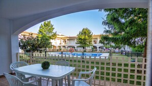 Un patio con una mesa y sillas blancas, una planta pequeña y vista a una piscina y edificios al fondo.