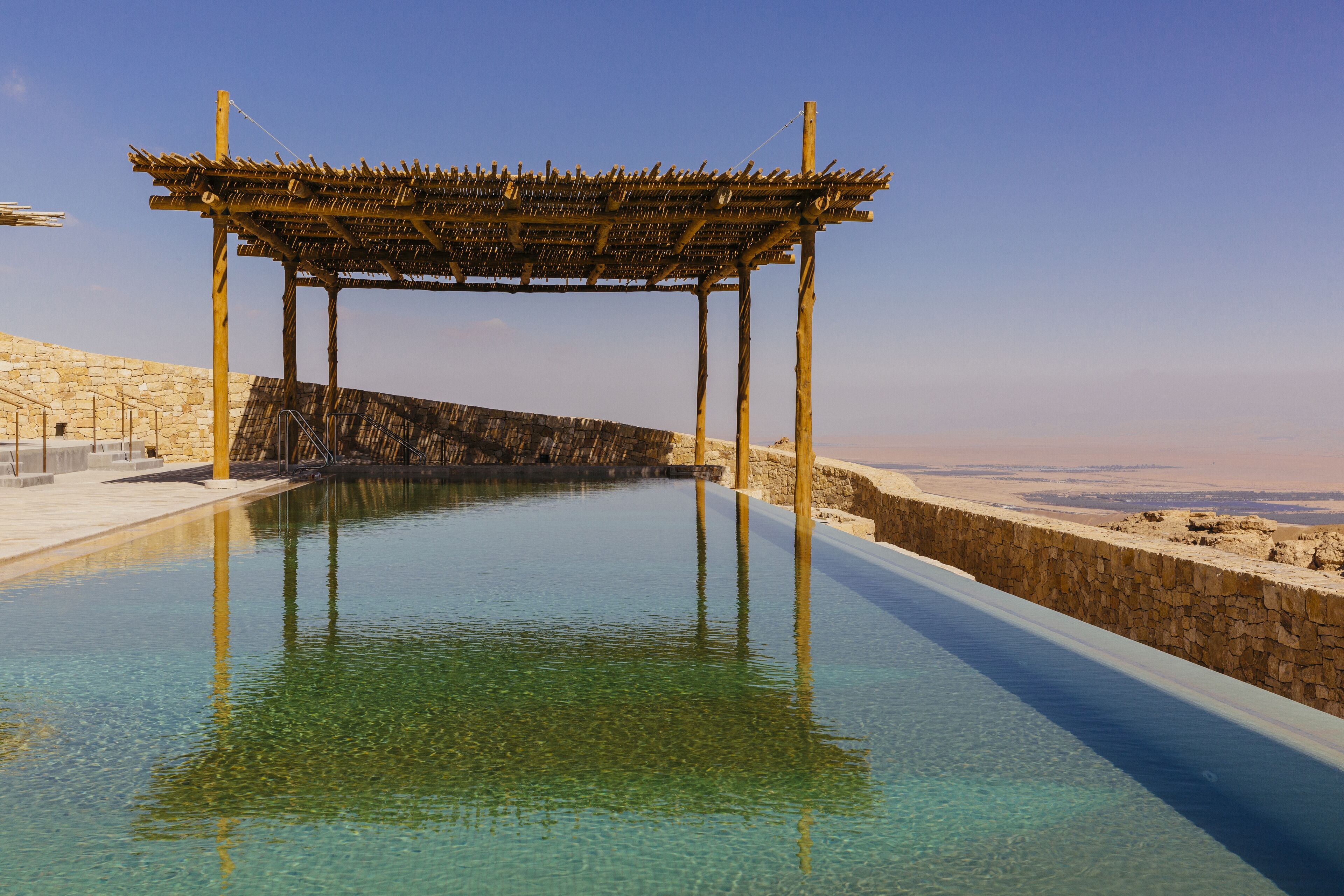 A clear pool with a wooden pergola structure, stone walls, and a view of the landscape.