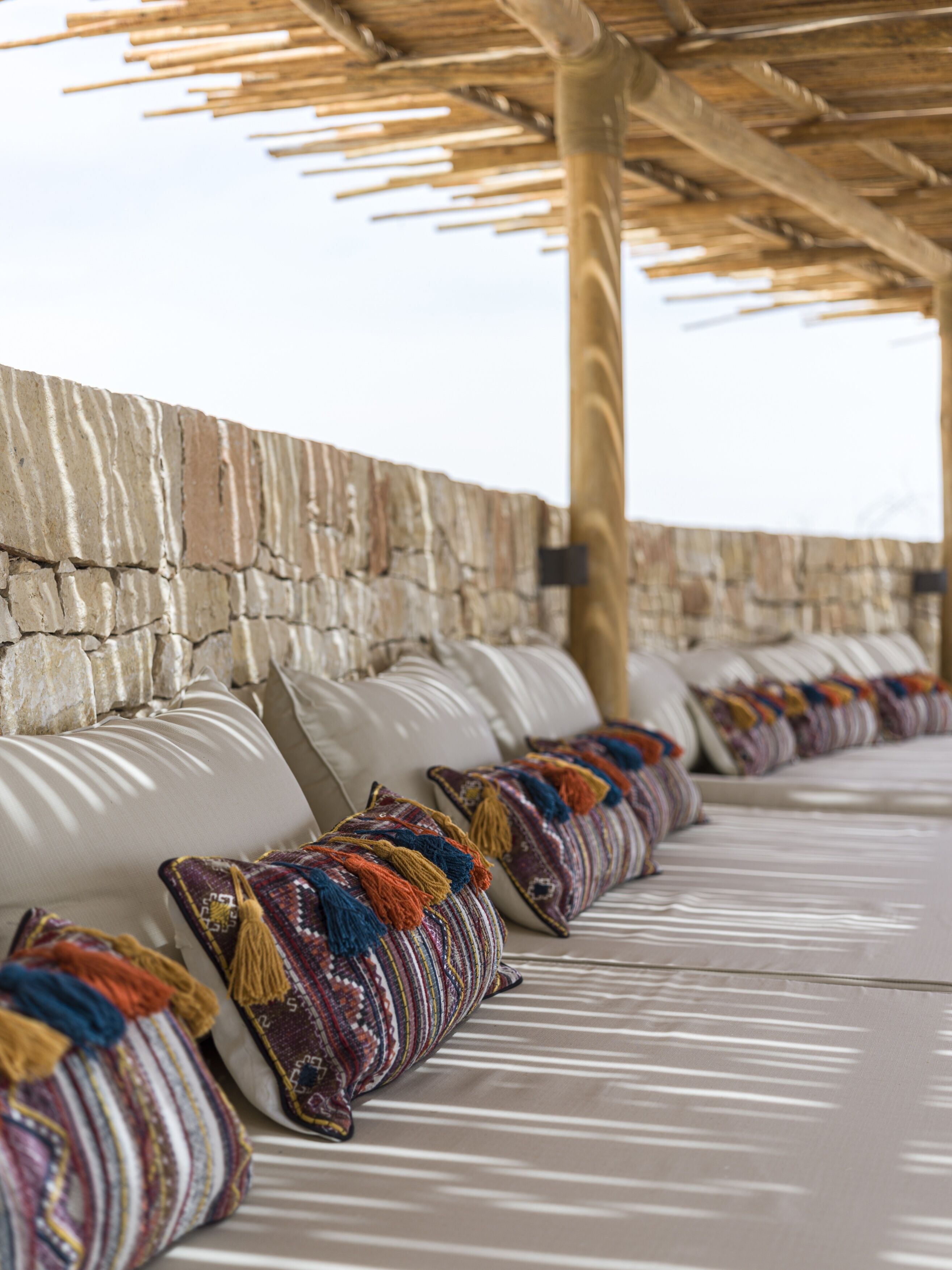 A row of white cushioned seating under a wooden pergola with a stone wall backdrop.