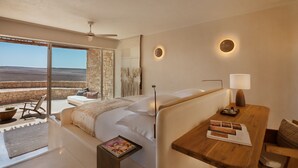 A modern hotel room with a large bed, a wooden side table, a reading area, and a balcony with a view of the landscape.