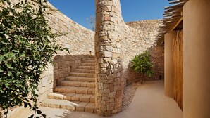 A stone-walled courtyard with a staircase leading to an upper level, surrounded by greenery and a wooden structure.