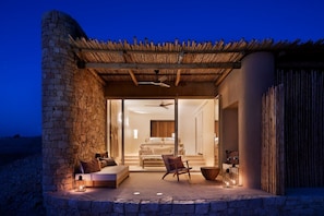 A stone-walled room with a glass door leading to a bedroom with a bed, a chair, and a table. - Six Senses Shaharut (Hevel Eilot)