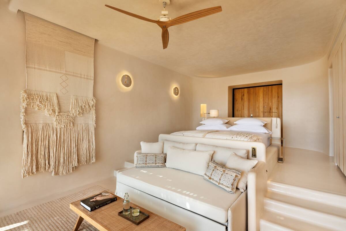 A modern bedroom with a white sofa, a ceiling fan, a large bed with white bedding, and a wall-mounted macrame piece.