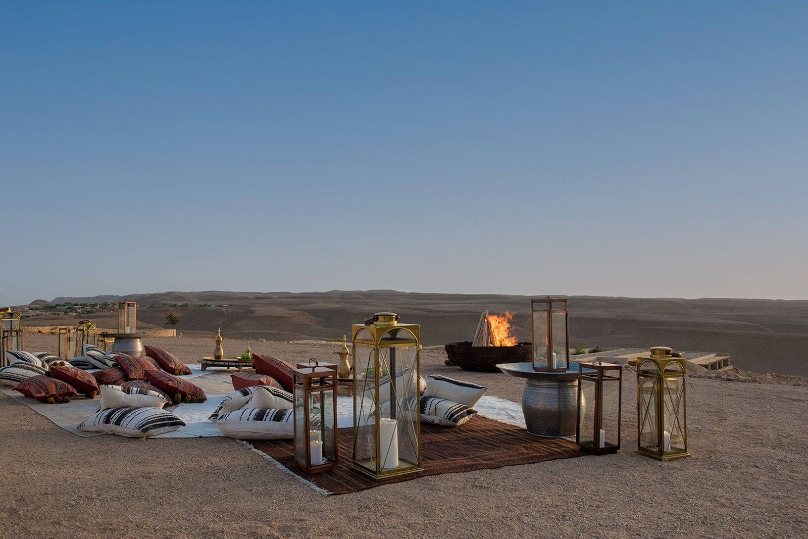 An outdoor seating area with cushions, a fire pit, and lanterns, set against a desert landscape.