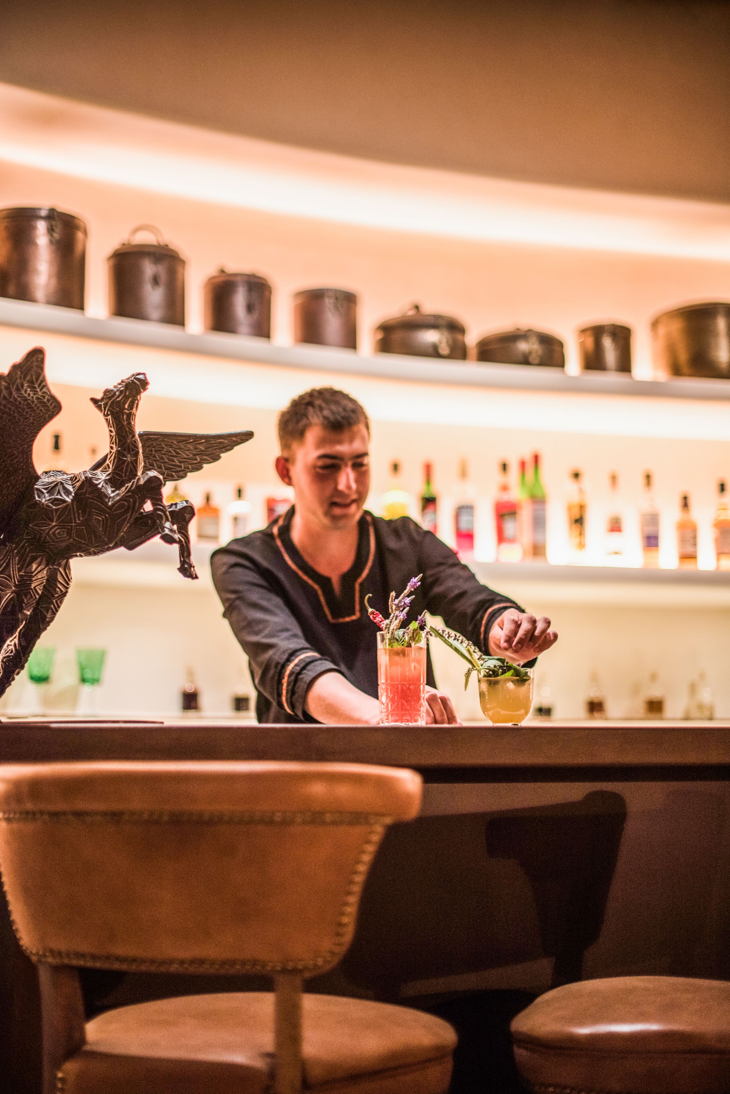 Zona de bar con un bartender preparando una bebida, estantes con botellas y una escultura metálica decorativa.