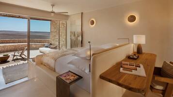 Un dormitorio moderno con una cama grande, una mesita de noche de madera, una lámpara de lectura y vistas al desierto a través de una puerta corrediza de vidrio.