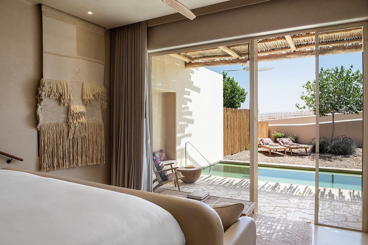 A modern hotel room with a large bed, a view of a swimming pool, and a patio area with lounge chairs.