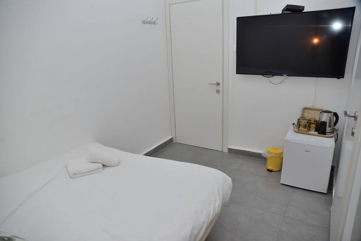 A small, clean room with a bed, a television, a small cabinet, and a trash bin.