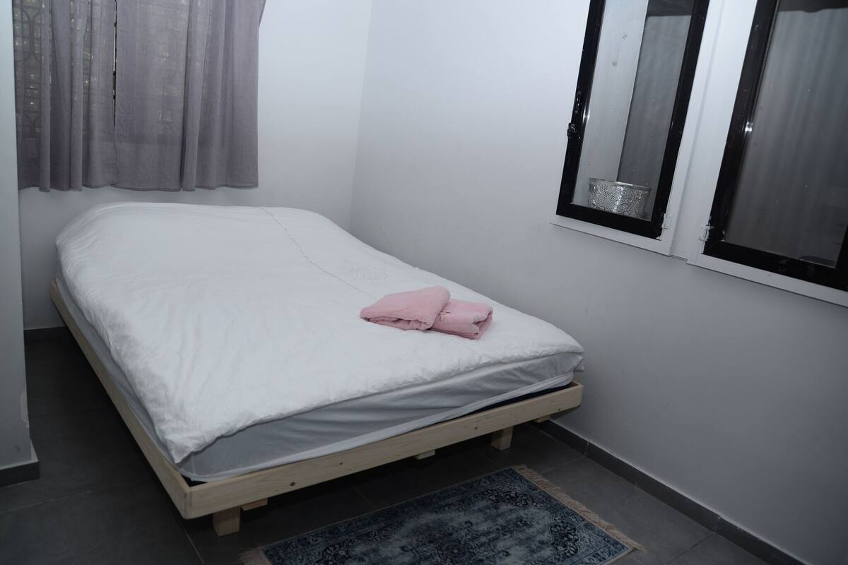 A single-bed room with a wooden frame, white bedding, a pink towel, a patterned rug, and a window with a metal basket on the sill.