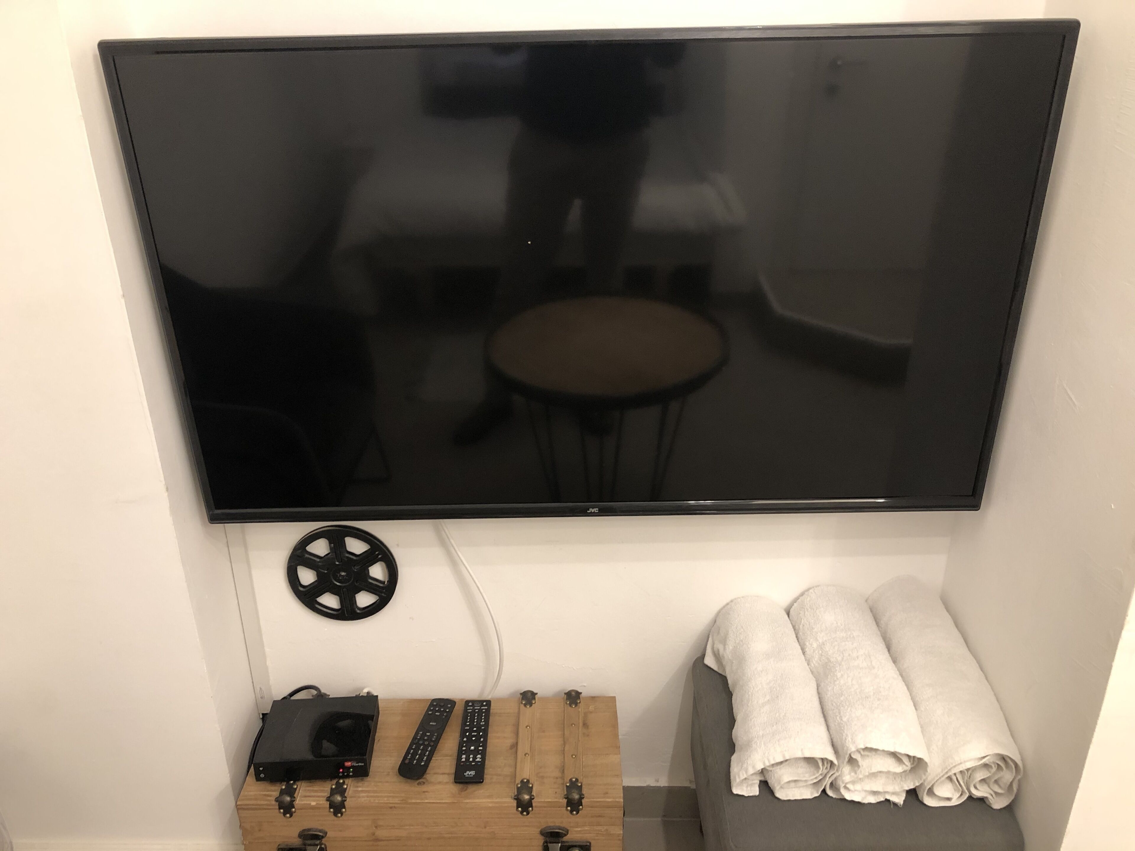 Un téléviseur à écran plat fixé au mur, une table en bois avec un projecteur et des télécommandes, et un banc avec des serviettes enroulées.