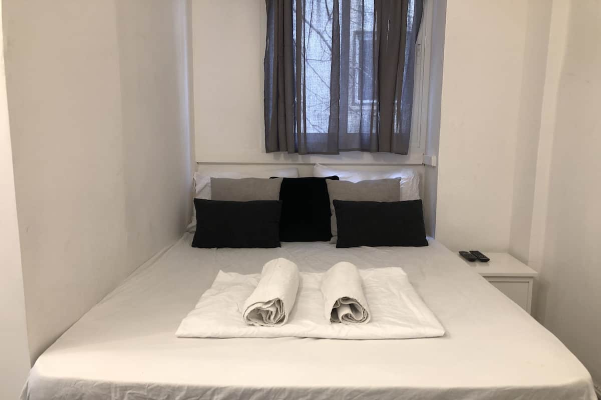 Ein ordentlich bezogenes Bett mit aufgerollten Handtüchern, ein Nachttisch mit Fernbedienung und ein Fenster mit Vorhängen.