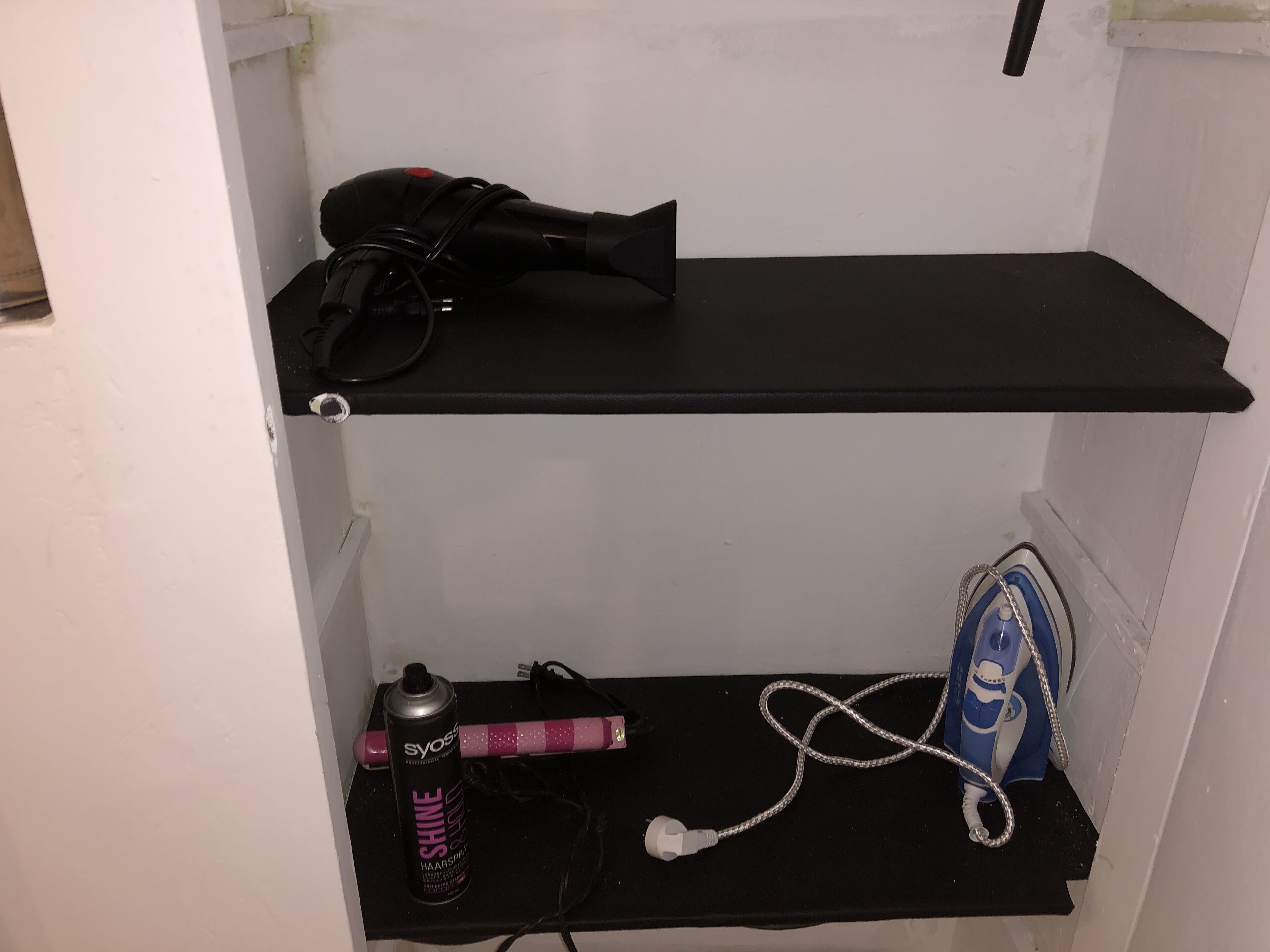 Un pequeño espacio de almacenamiento con un estante negro que contiene un secador de pelo, un bote de laca y un cepillo azul.