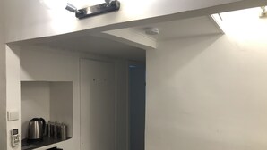 Un pasillo estrecho con una puerta blanca a la izquierda, un refrigerador negro y una estantería con utensilios de cocina.