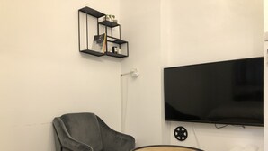 Una sala de estar con un sillón gris, una mesita redonda pequeña, un televisor montado en la pared y un estante negro con objetos decorativos.