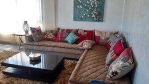 A modern living room with a sectional sofa, a glass coffee table, and a floral painting on the wall. - Residence Galets Sur Mer 4701 (Dar Bouazza)