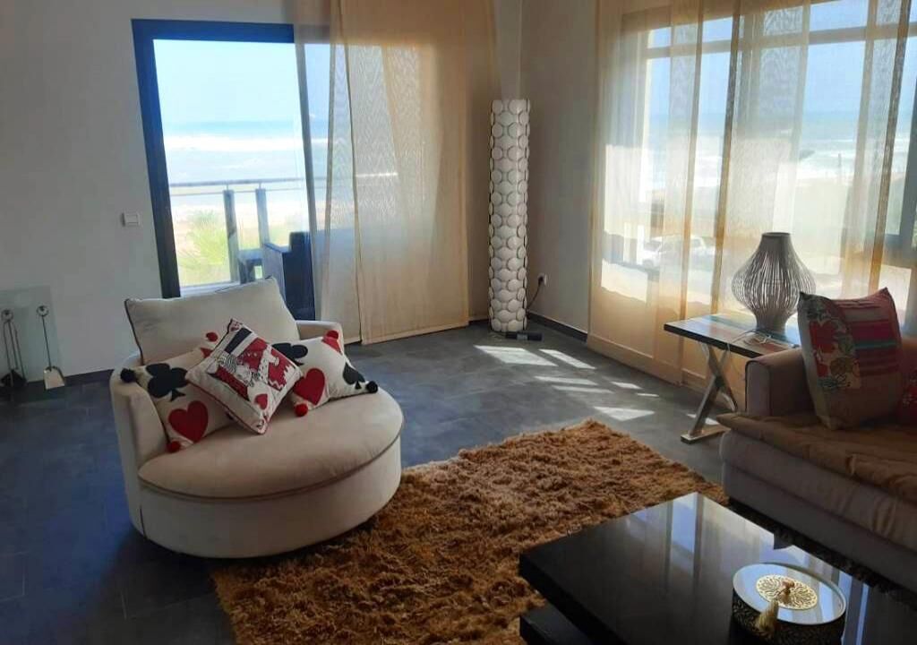 Een moderne woonkamer met een groot raam, een ronde witte bank met opvallende kussens, een bruin vloerkleed en een klein bijzettafeltje met een lamp.