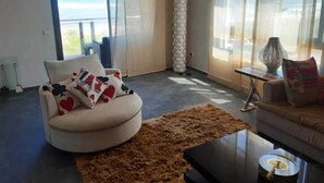 A modern living room with a large window, a round white sofa with patterned cushions, a brown rug, and a small side table with a lamp. - Residence Galets Sur Mer 4701 (Dar Bouazza)