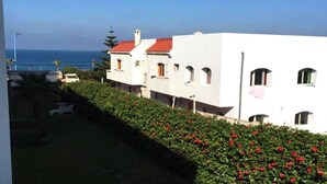 Eine Reihe weißer Gebäude mit roten Dächern, ein gepflegter Heckenstreifen mit roten Blüten und der Blick auf das Meer.